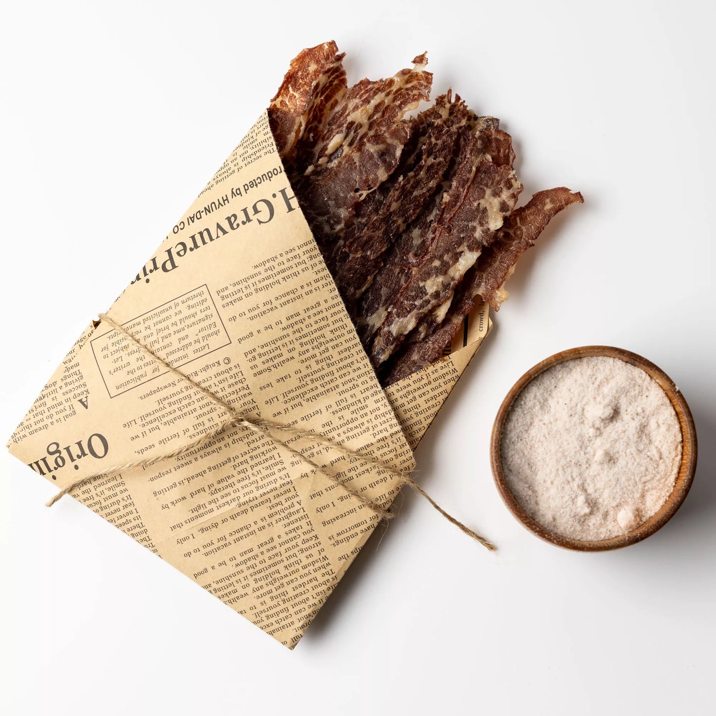 Dried meat slices in a newspaper-wrapped package with a bowl of seasoning on a white background
