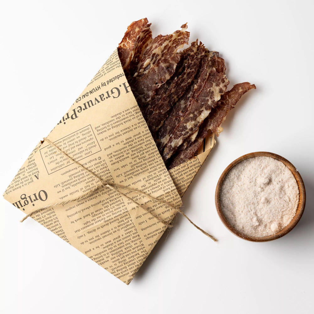 
                  
                    Dried meat slices in a newspaper-wrapped package with a bowl of seasoning on a white background
                  
                